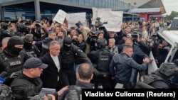 Vlad Plahotniuc escortat spre microbuzul care armează să-l transporte la penitenciarul 13. În plan secund - câteva persoane țin pancarde în susținerea acestuia. Aeroportul Internațional Chișinău, 25 septembrie 